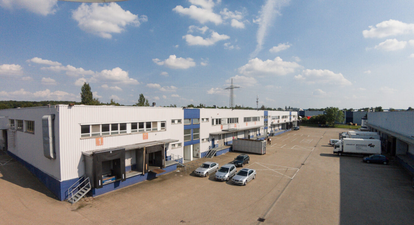 Ein großes Industrielager mit mehreren Laderampen, geparkten Autos und Lieferwagen auf einem großen, gepflasterten Parkplatz an einem teilweise bewölkten Tag.