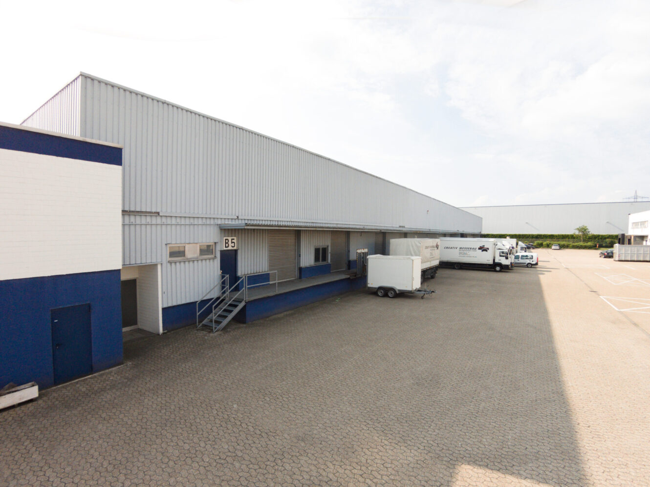 Außenansicht eines großen Industrielagers mit einer Laderampe, mehreren Anhängern und einem Lkw, der auf einem gepflasterten Grundstück bei teilweise bewölktem Himmel abgestellt ist.