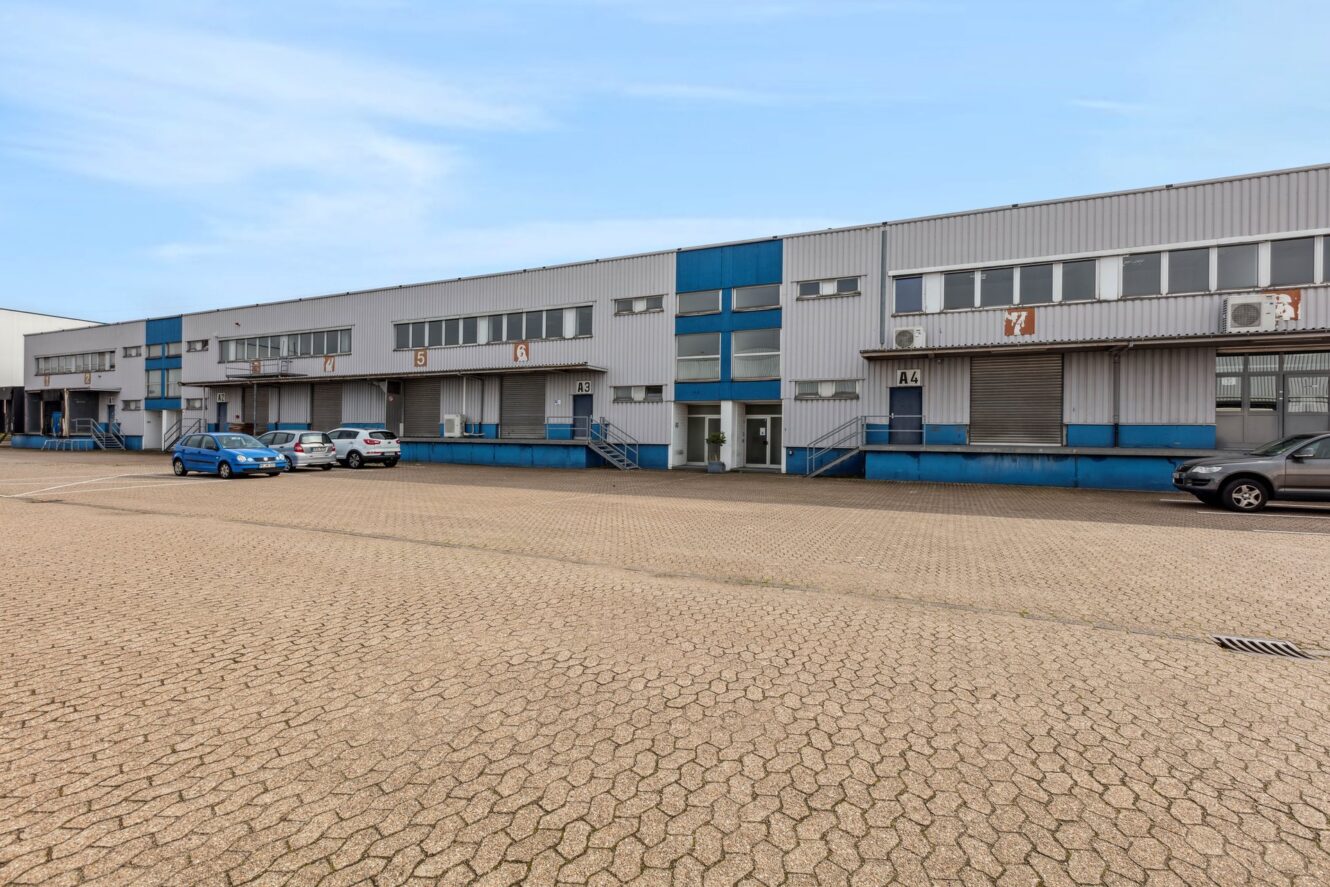 Ein großes Industrielager mit mehreren Laderampen, ein paar geparkten Autos und einem gepflasterten Parkplatz davor. Das Gebäude ist grau mit blauen Akzenten und hat nummerierte Buchten.