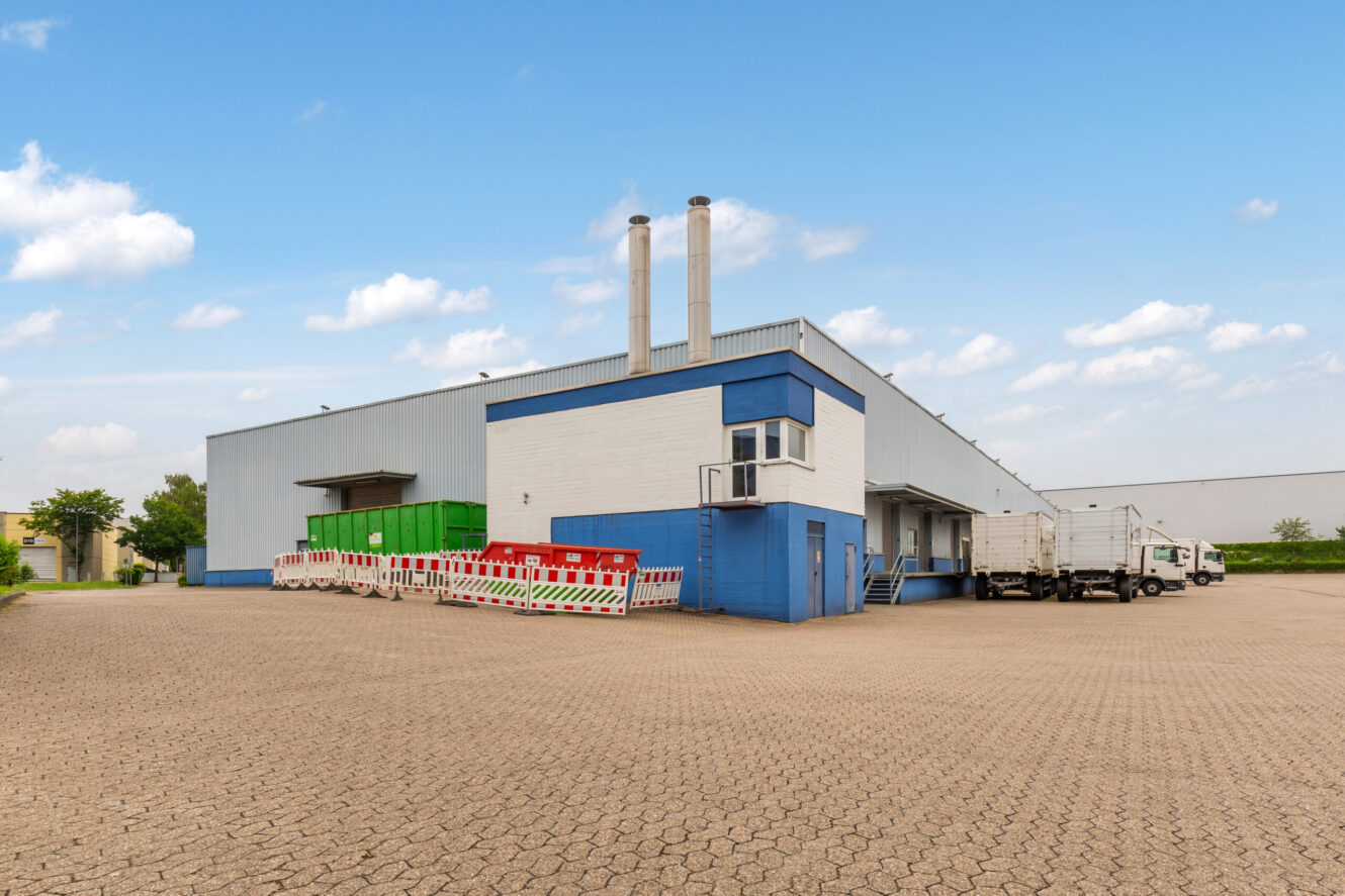 Ein großes Industrielager mit Laderampen, grünen Containern, roten Müllcontainern und mehreren weißen Lastwagen, die unter blauem Himmel auf einem gepflasterten Platz geparkt sind.