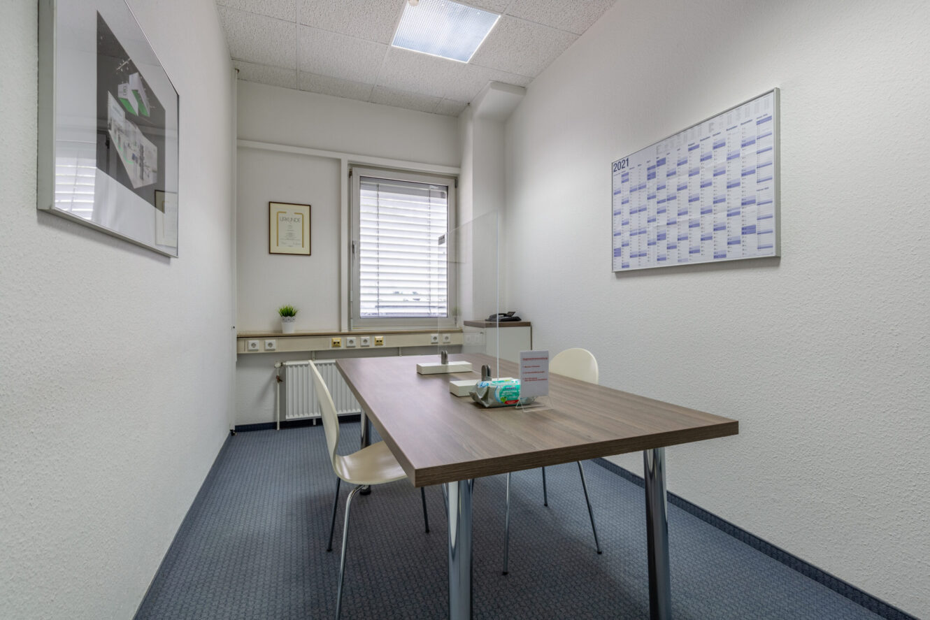 Kleines Büro mit einem Holztisch, zwei Stühlen, einem Wandkalender, einem gerahmten Bild, einem Fenster mit Jalousien und minimaler Ausstattung.