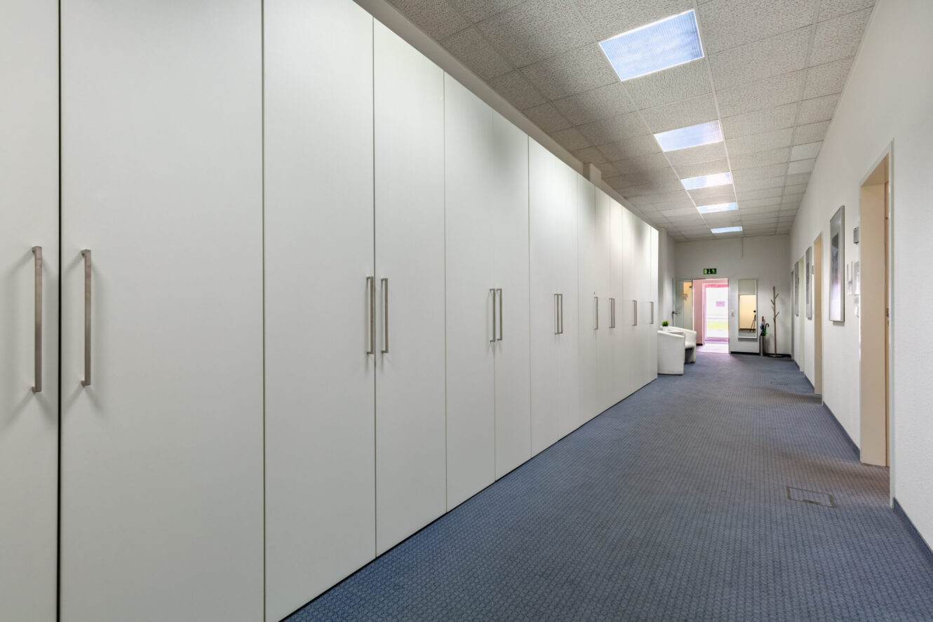 Ein langer Flur mit weißen Einbauschränken auf einer Seite, blauem Teppichboden und Deckenleuchten. Der Flur führt zu einem Sitzbereich in der Nähe einer gläsernen Ausgangstür.