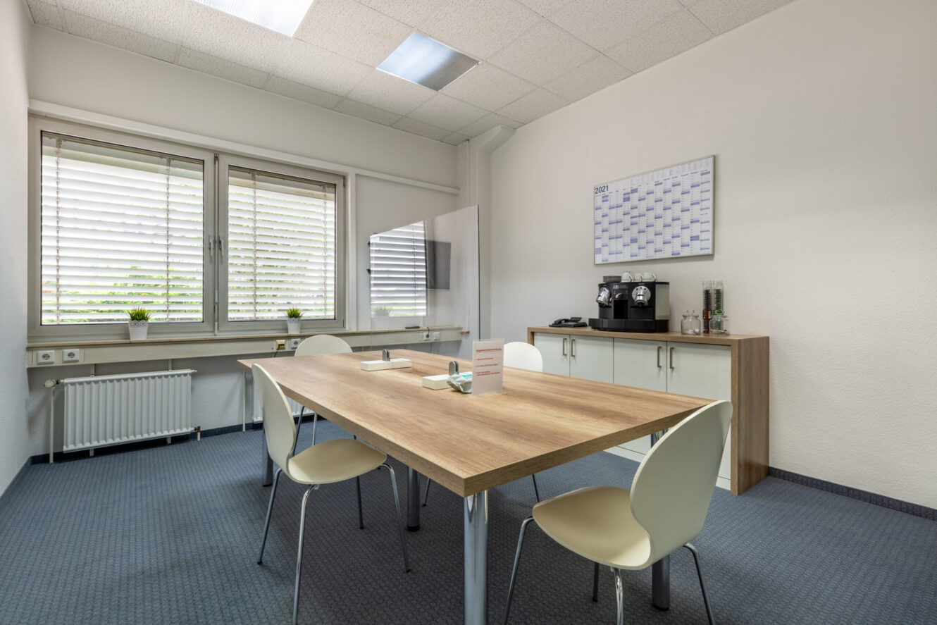 Ein kleiner Besprechungsraum mit einem Holztisch, vier weißen Stühlen, einem Wandkalender, einer Kaffeemaschine und großen Fenstern mit Jalousien.