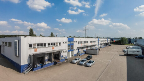 Ein großes Industrielager mit Laderampen, geparkten Autos und einem Lkw auf dem Parkplatz bei teilweise bewölktem Himmel.