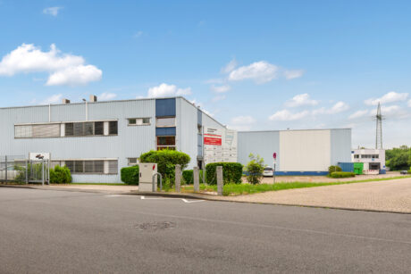 Ein modernes Industriegebäude mit Metallverkleidung, ein Parkplatz und ein großes Schild stehen unter einem blauen Himmel mit vereinzelten Wolken.