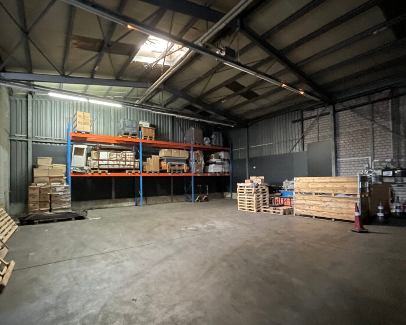 Ein Lagerhaus mit Metallregalen, in denen Kisten und Paletten stehen, die teilweise an der Wand und auf dem Boden gestapelt sind, unter einem hohen Wellblechdach.