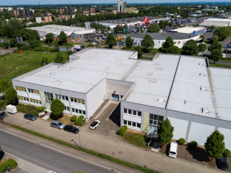 Luftaufnahme eines großen industriellen oder gewerblichen Gebäudekomplexes mit weißen Dächern, angrenzenden Parkplätzen und umliegenden Grünflächen in einem städtischen Gebiet.