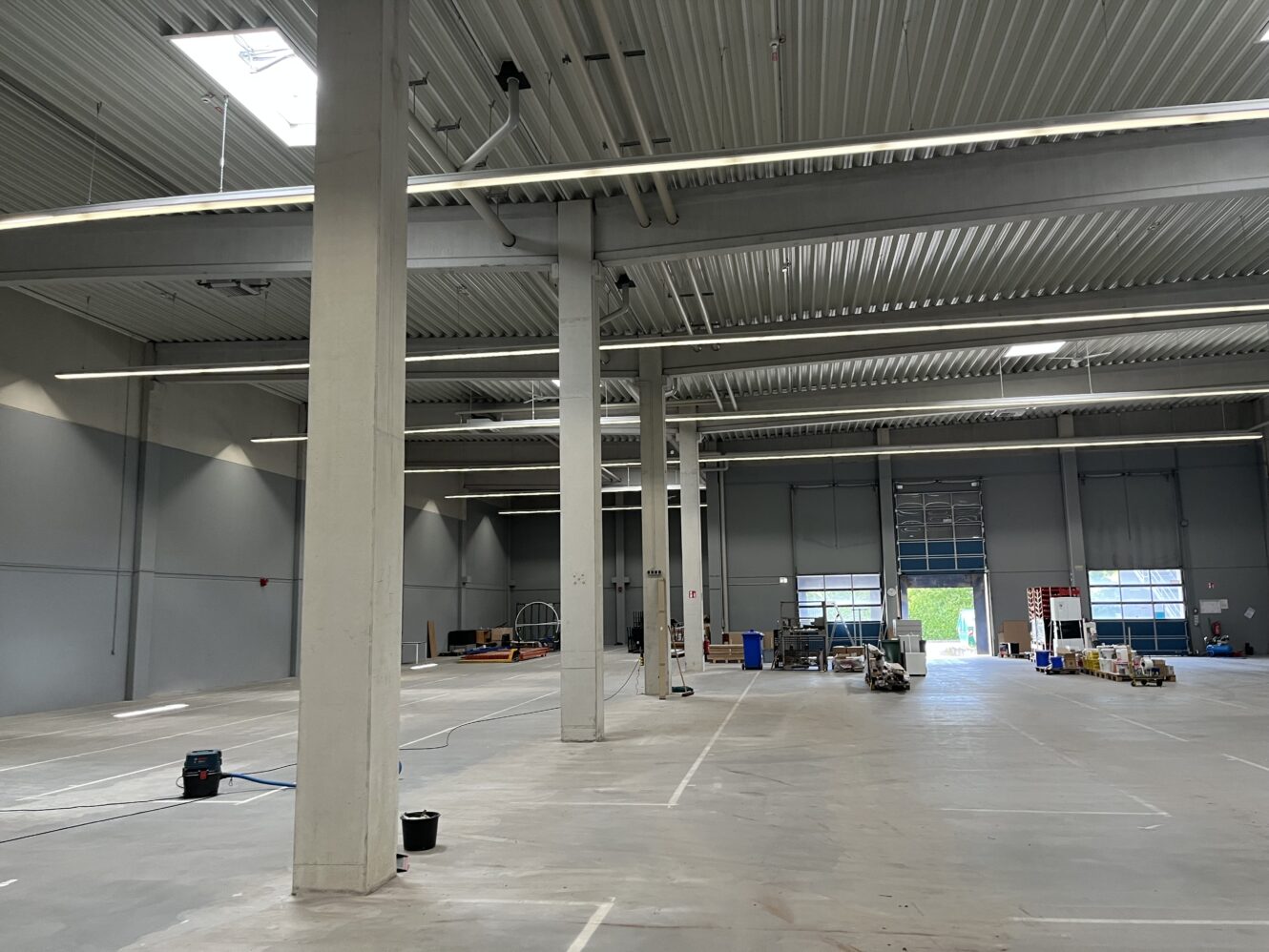 Ein geräumiges, größtenteils leeres Lagerhaus mit freiliegenden Balken, verstreuten Geräten, gemalten Linien auf dem Boden und einem offenen Garagentor, das Tageslicht hereinlässt.