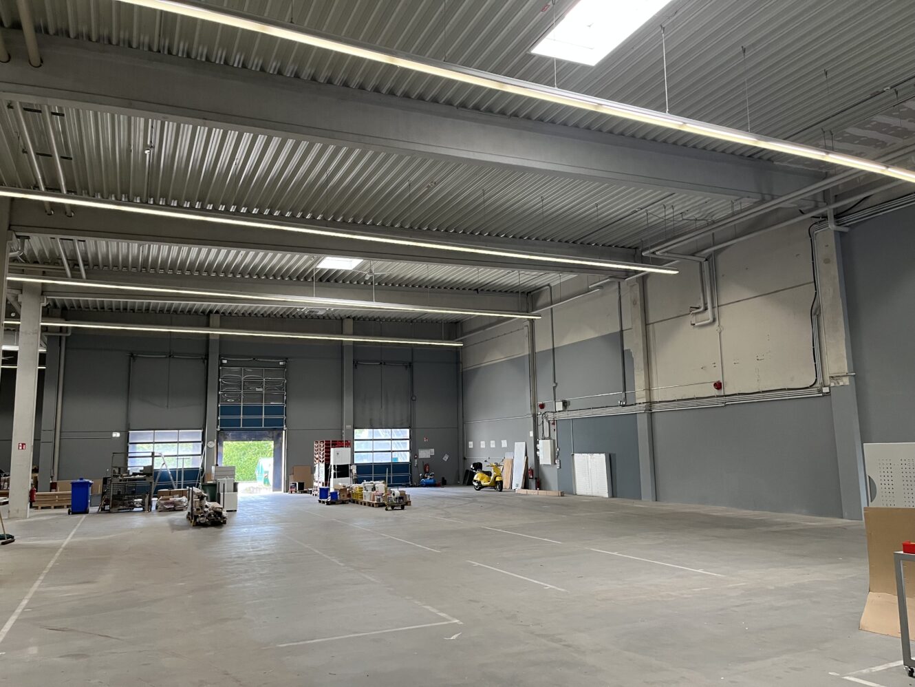 Große, größtenteils leere Lagerhalle mit hohen Decken, grauen Wänden und verstreuten Geräten und Kisten im hinteren Bereich. Natürliches Licht fällt durch Fenster und ein offenes Garagentor ein.