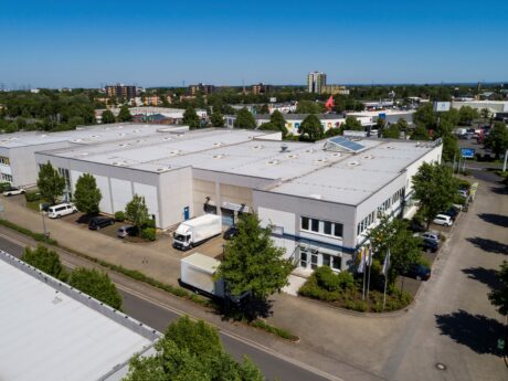 Luftaufnahme eines großen, weißen Industrielagers mit davor geparkten Lastwagen, umgeben von Bäumen und Straßen in einem Gewerbegebiet.