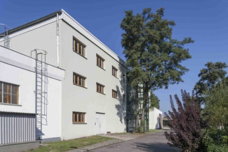 Ein weißes, dreistöckiges Industriegebäude mit holzgerahmten Fenstern und einer Außentreppe aus Metall steht neben einem Baum unter einem strahlend blauen Himmel.