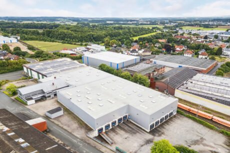 Luftaufnahme eines industriellen Lagerkomplexes mit mehreren Laderampen, umgeben von Straßen, Grünflächen und nahe gelegenen Gebäuden in einem halb-ländlichen Gebiet.