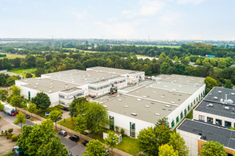 Luftaufnahme eines großen industriellen oder gewerblichen Gebäudekomplexes mit weißen Dächern, umgeben von Bäumen, Straßen und grünen Feldern unter einem teilweise bewölkten Himmel.