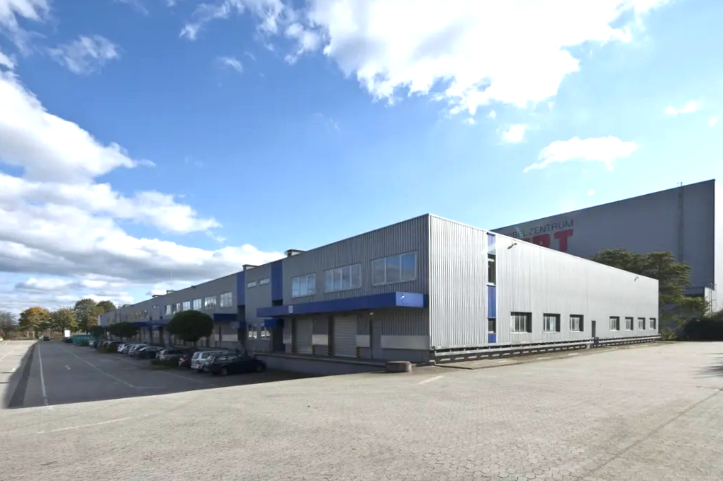 Große Industriehalle mit Metallverkleidung und blauen Akzenten, umgeben von einem leeren Parkplatz unter einem teilweise bewölkten Himmel.