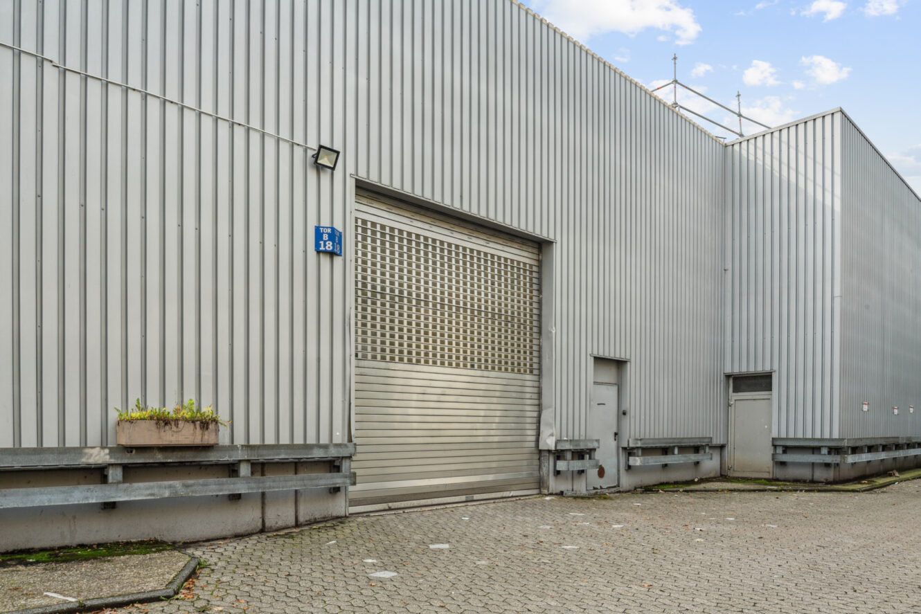 Großes Industriegebäude mit Wellblechverkleidung, einem Hauptrolltor, zwei kleineren Toren, einem Blumenkasten und gepflastertem Boden unter einem teilweise bewölkten Himmel.