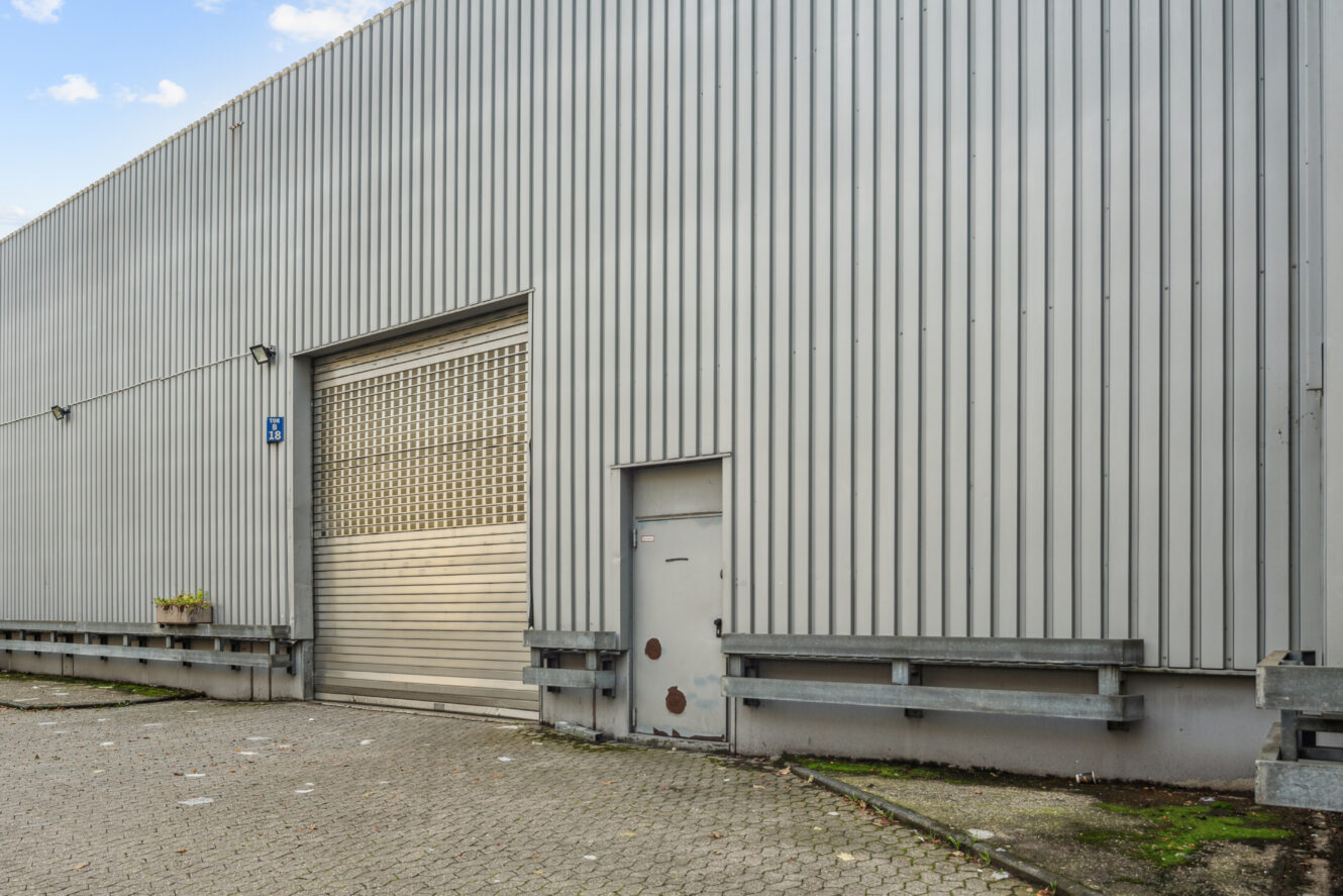 Außenansicht eines grauen Industrielagers mit einem großen Rolltor aus Metall, einem kleineren Zugangstor und einer gepflasterten Fläche davor.