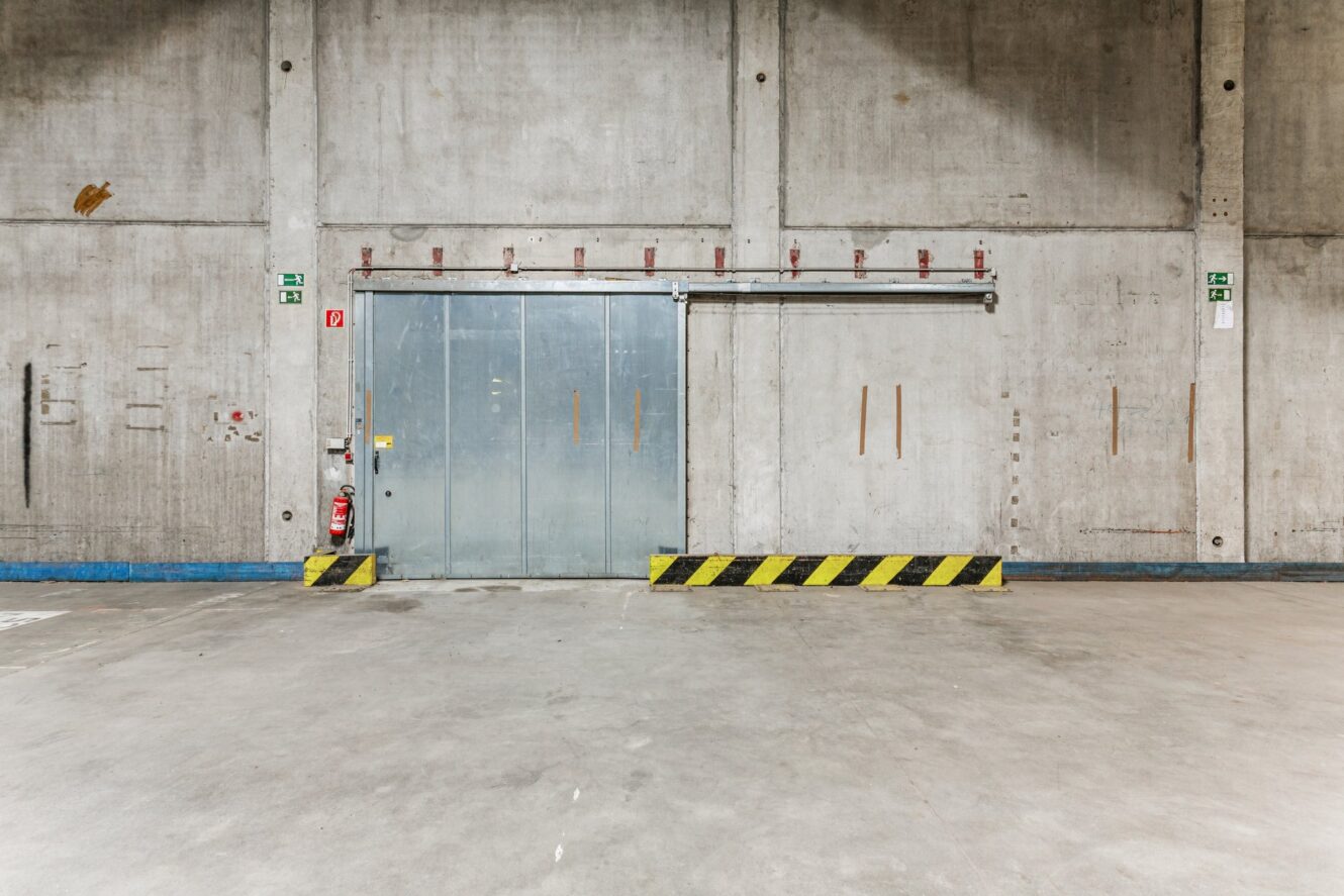 Große Metallschiebetür in einer Betonwand eines leeren Industrielagers, mit einer gelb-schwarz gestreiften Absperrung und einem Feuerlöscher auf der linken Seite.