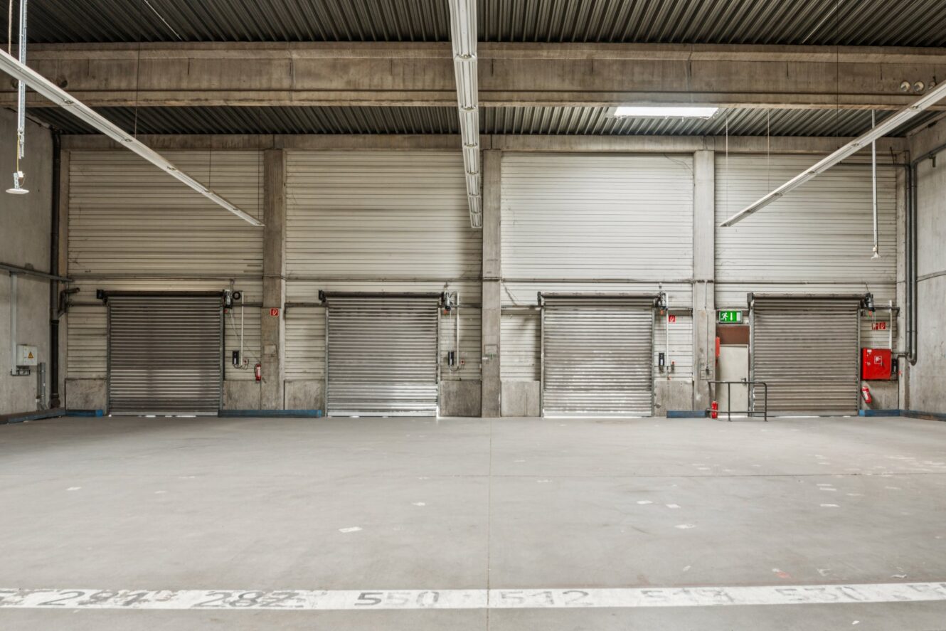 Innenraum einer leeren Lagerhalle mit vier geschlossenen Metallrolltoren, Betonboden und sichtbarer Brandschutzausrüstung an der rechten Wand.