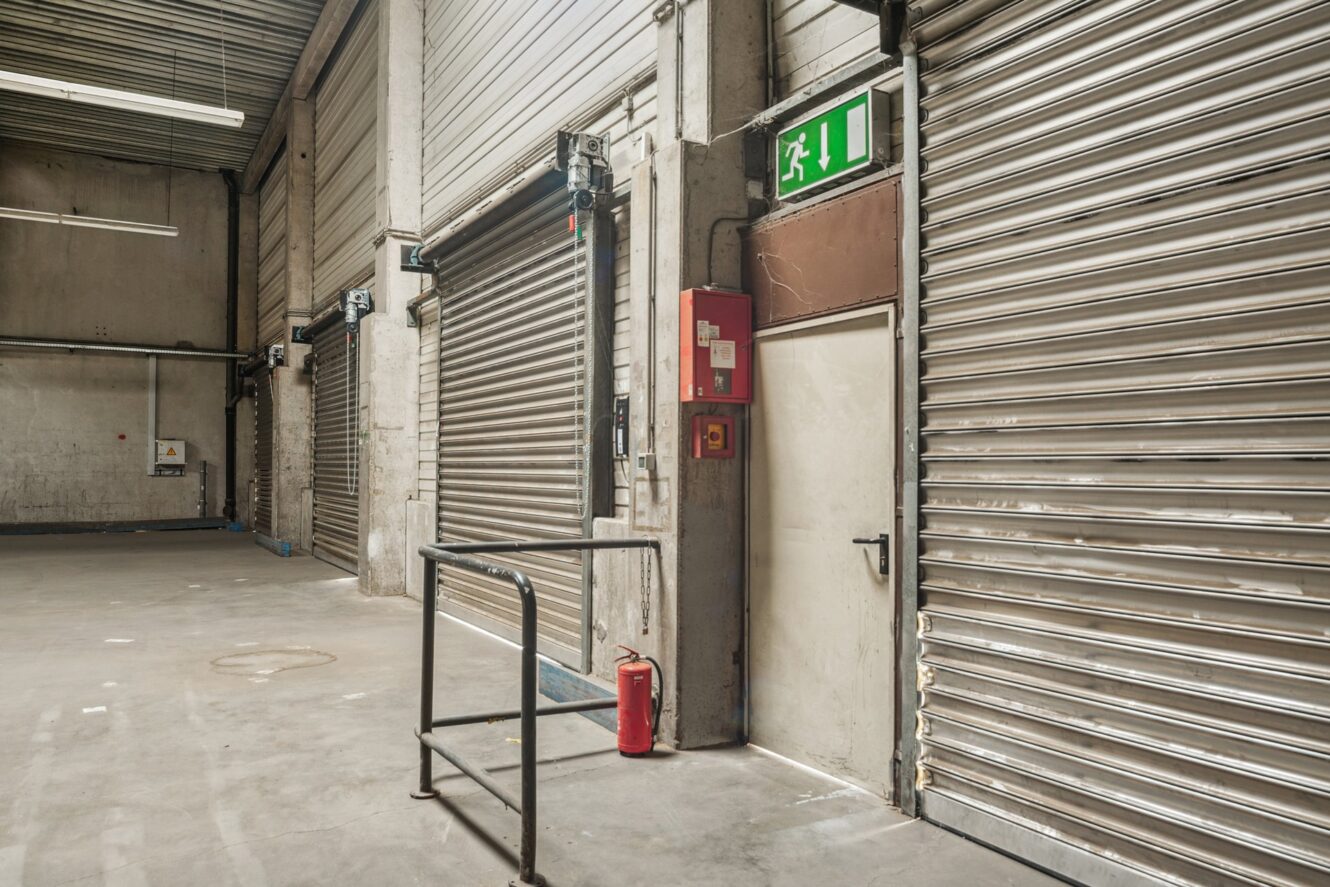 Leeres Industrielager mit geschlossenen Metallrollläden, einem Feuerlöscher, einem Notausgangsschild und an der Wand montierter Sicherheitsausrüstung.