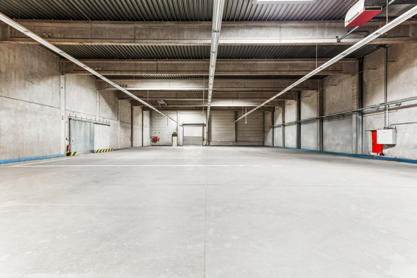 Leeres Industrielager mit Betonböden, hohen Decken, freiliegenden Balken und großen geschlossenen Garagentoren im hinteren Bereich.