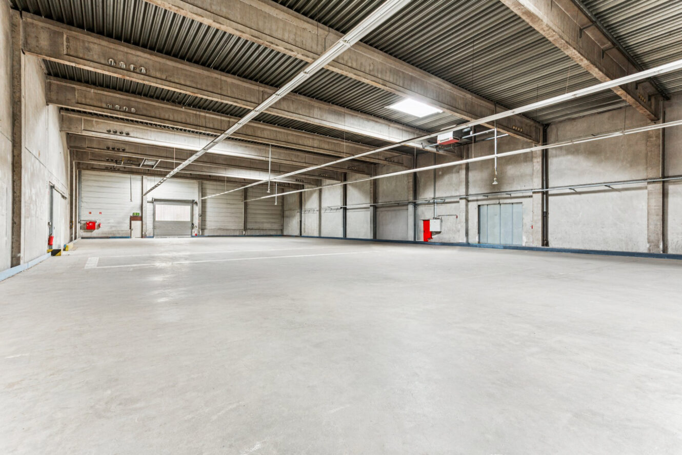 Weitwinkelansicht eines leeren Industrielagers mit Betonböden, hohen Decken, freiliegenden Balken und Leuchtstoffröhrenbeleuchtung. An den Wänden sind Feuerlöscher angebracht.