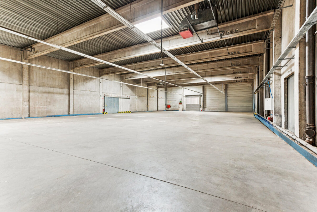 Geräumiges, leeres Industrielager mit hohen Decken, Sichtbetonwänden, großen Rolltoren und Deckenbeleuchtung.