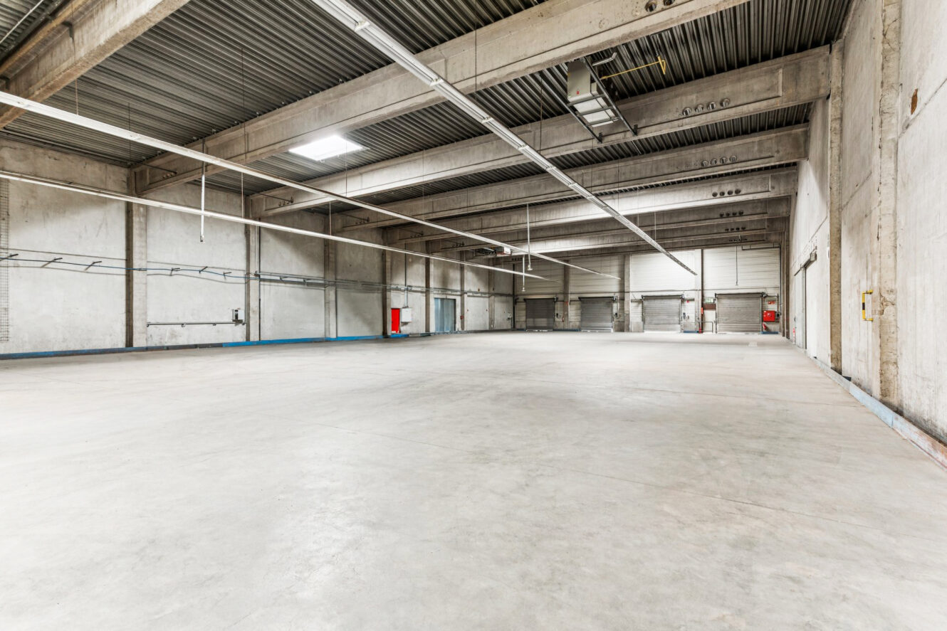 Große, leere Industrielagerhalle mit Betonböden, freiliegenden Stahlträgern, heller Deckenbeleuchtung und an den Wänden angebrachten Brandschutzvorrichtungen.