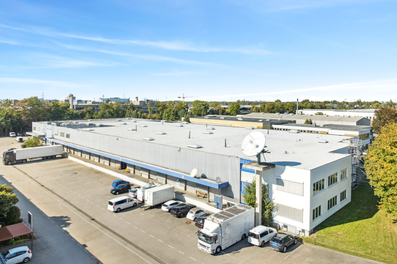 Großes weißes Industrielager mit mehreren Ladebuchten, geparkten Fahrzeugen und einer Satellitenschüssel auf dem Dach, umgeben von Bäumen und unter freiem Himmel.