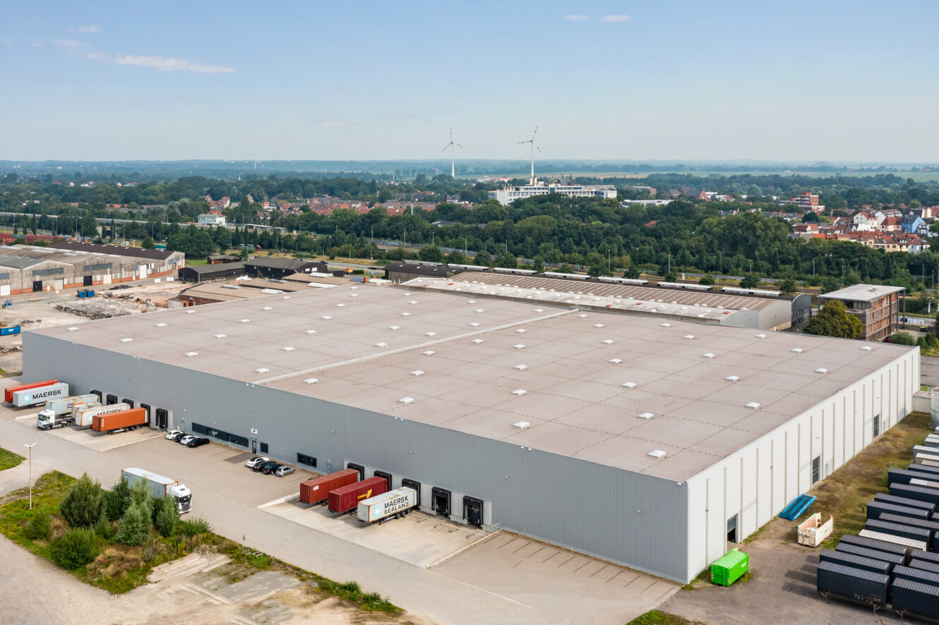 Luftaufnahme eines großen Industrielagers mit mehreren Laderampen und draußen geparkten Lastwagen, umgeben von anderen Industriegebäuden und Grünanlagen.