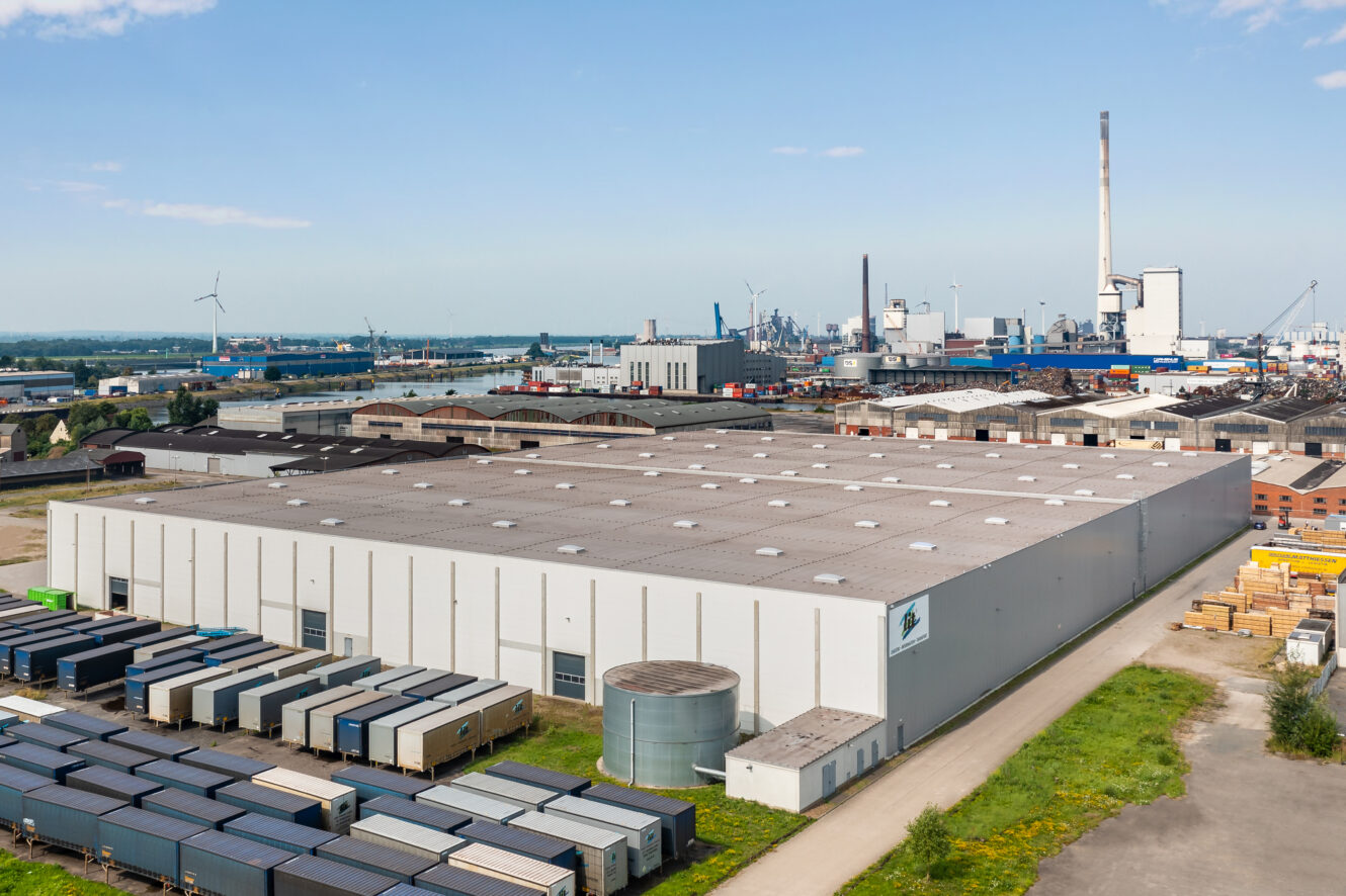Im Vordergrund steht ein großes weißes Industrielager, umgeben von Schiffscontainern, während im Hintergrund unter klarem Himmel Fabriken und Schornsteine zu sehen sind.