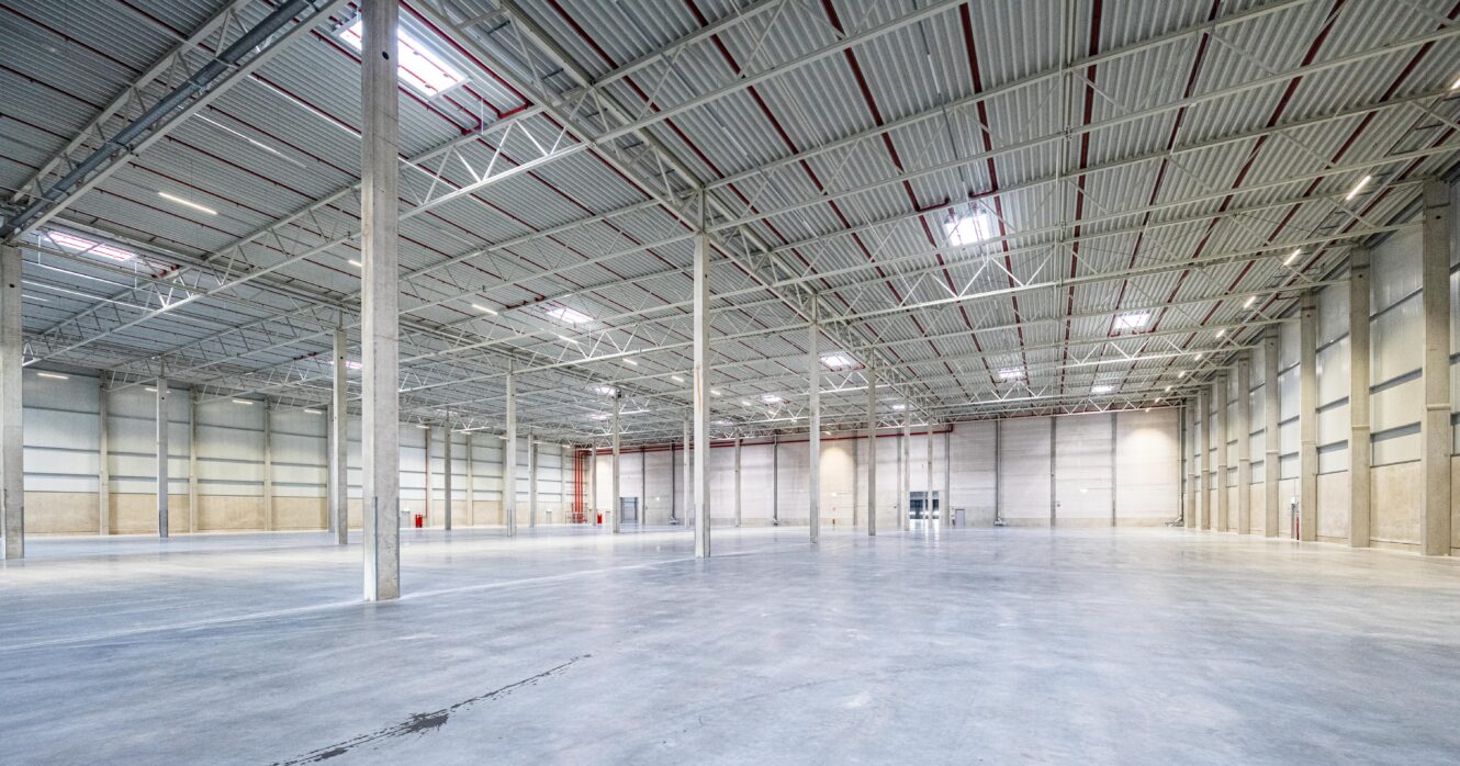 Großes, leeres Lagerhaus mit Betonboden, hoher Decke, Metallträgern, Stützpfeilern und Oberlichtern, die Tageslicht hereinlassen.