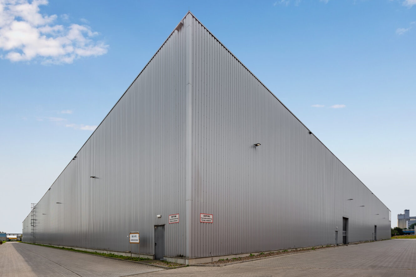 Ein großes, silbernes, dreieckiges Industrielager mit Metallverkleidung, Sicherheitskameras und Schildern an der Außenseite, vor einem klaren blauen Himmel.