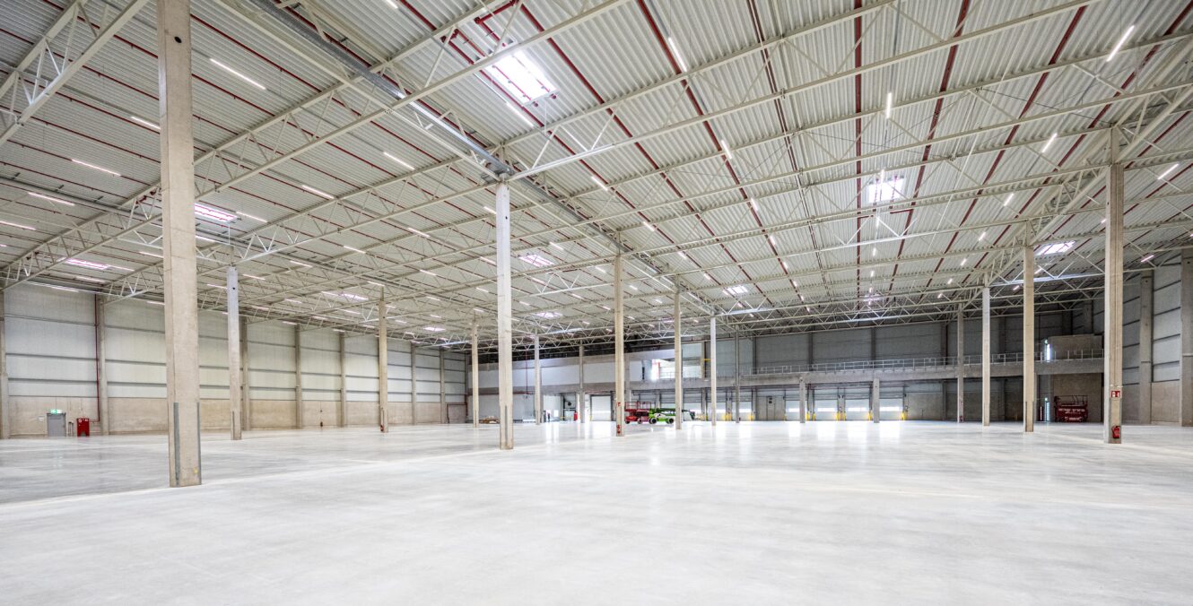 Geräumige, leere Lagerhalle mit hohen Decken, Betonböden, Stützpfeilern und Deckenbeleuchtung. Ein roter Gabelstapler ist an der Rückwand geparkt.