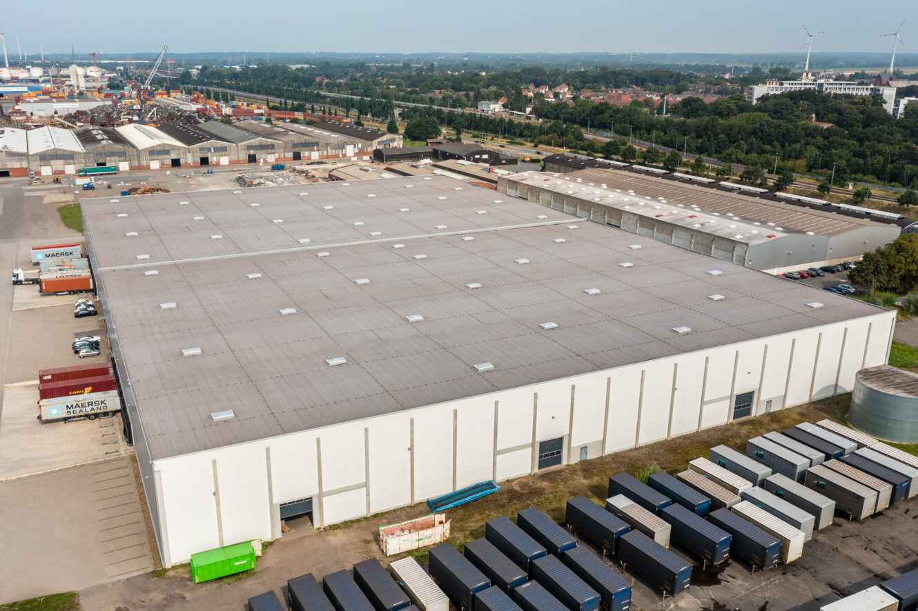 Luftaufnahme eines großen Industrielagers mit mehreren Laderampen und im Freien geparkten Lastwagen in einem Industriegebiet.
