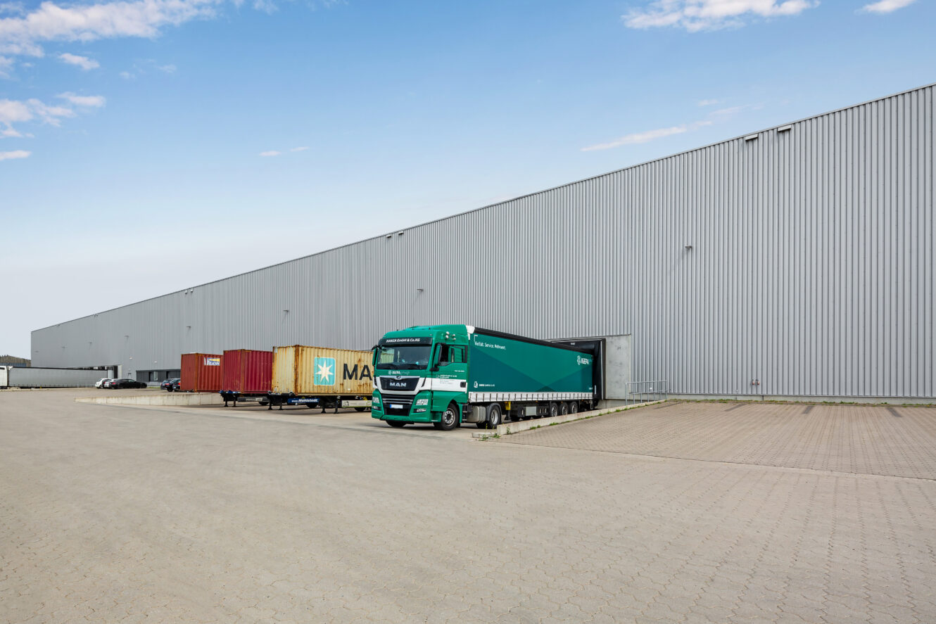 Ein grüner Sattelschlepper fährt rückwärts an eine Laderampe in einem großen Industrielager heran, in dessen Nähe sich unter blauem Himmel mehrere Frachtcontainer befinden.
