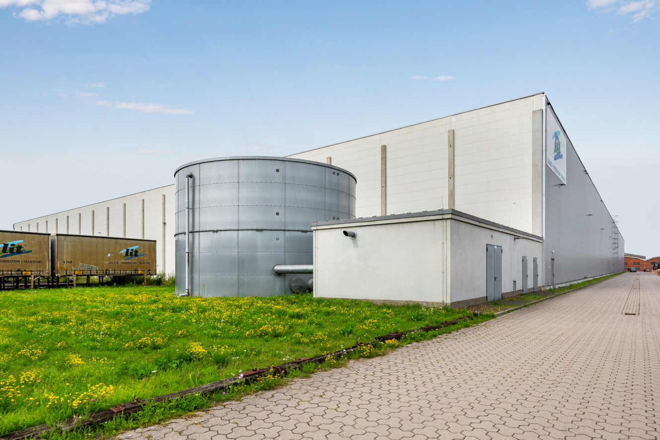 Großes Industrielager mit Metalltank und kleinem Wirtschaftsgebäude auf einer Grünfläche, angrenzend an einen gepflasterten Gehweg unter freiem Himmel.
