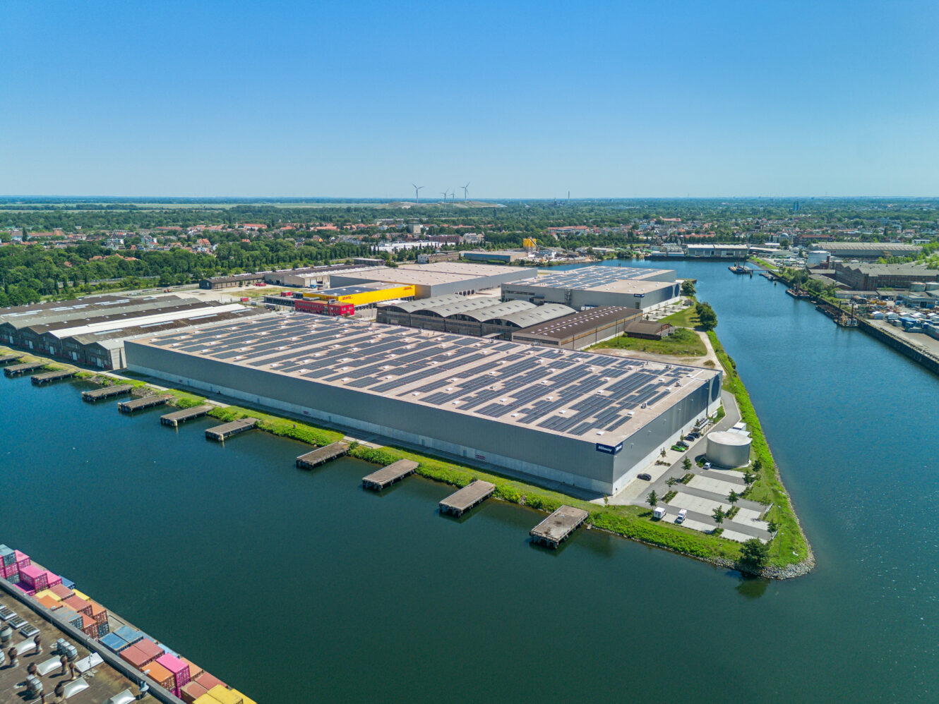 Luftaufnahme eines großen Industrielagers mit Solarzellen auf dem Dach, das an einer Wasserstraße liegt, mit Schiffsdocks und einer Stadtlandschaft im Hintergrund.
