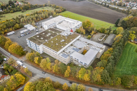 Luftaufnahme eines großen Industrie- oder Bürokomplexes, umgeben von Bäumen, Feldern und einem Parkplatz mit Lastwagen und Autos.