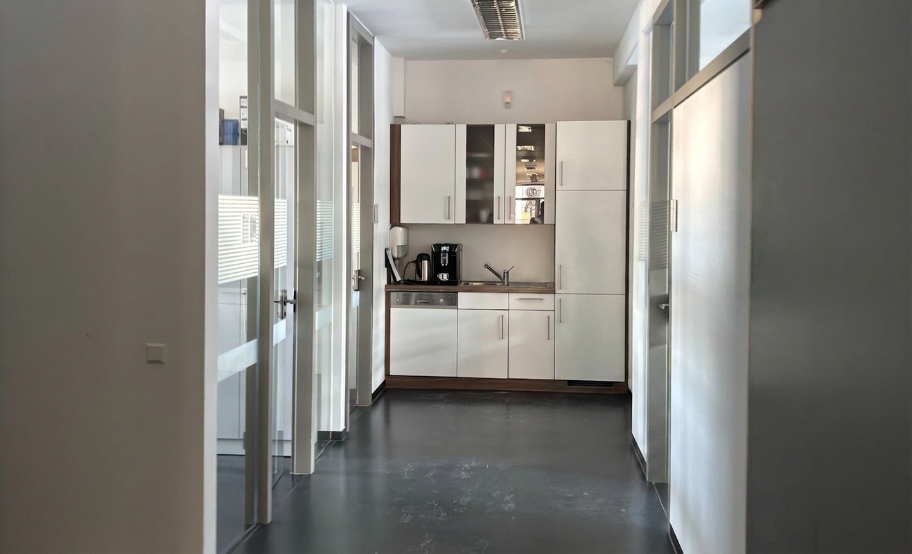 Ein moderner Flur führt zu einer kleinen Küchenzeile mit weißen Schränken, einer Kaffeemaschine und einem Waschbecken; links befinden sich Glastüren.