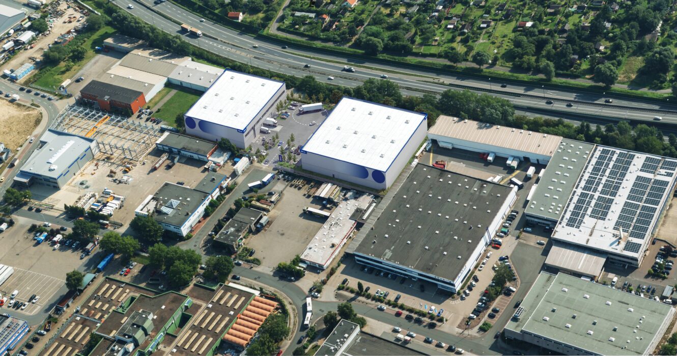 Luftaufnahme eines Industriekomplexes mit Lagerhallen, angrenzenden Gebäuden, Parkplätzen, einer Autobahn und Bäumen mit Häusern in der Nähe.