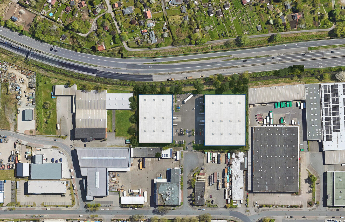 Luftaufnahme eines Industriekomplexes mit Lagerhallen, Parkplätzen, Straßen, Grünanlagen, einer Autobahn und Wohngrundstücken.