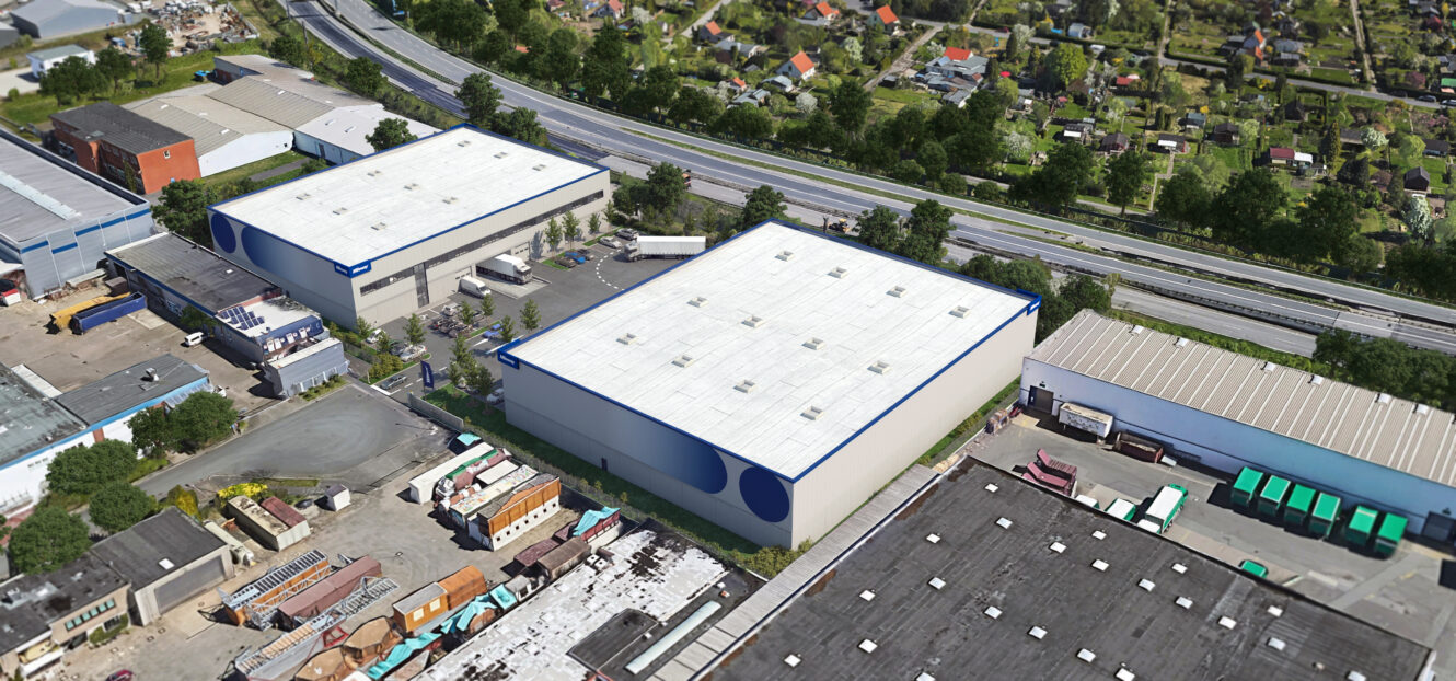 Luftaufnahme von zwei großen Lagerhäusern mit weißen Dächern, umgeben von Straßen, Lastwagen und nahe gelegenen Industrie- und Wohngebieten.