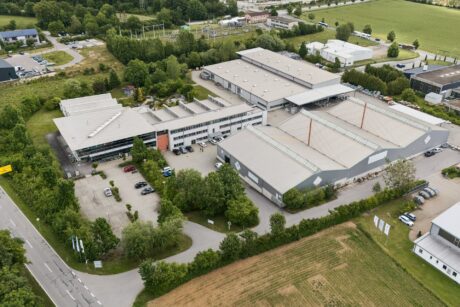 Luftaufnahme eines Industriekomplexes mit mehreren großen Lagerhallen, Parkplätzen, Bäumen und den umliegenden Straßen in einer halb ländlichen Gegend.