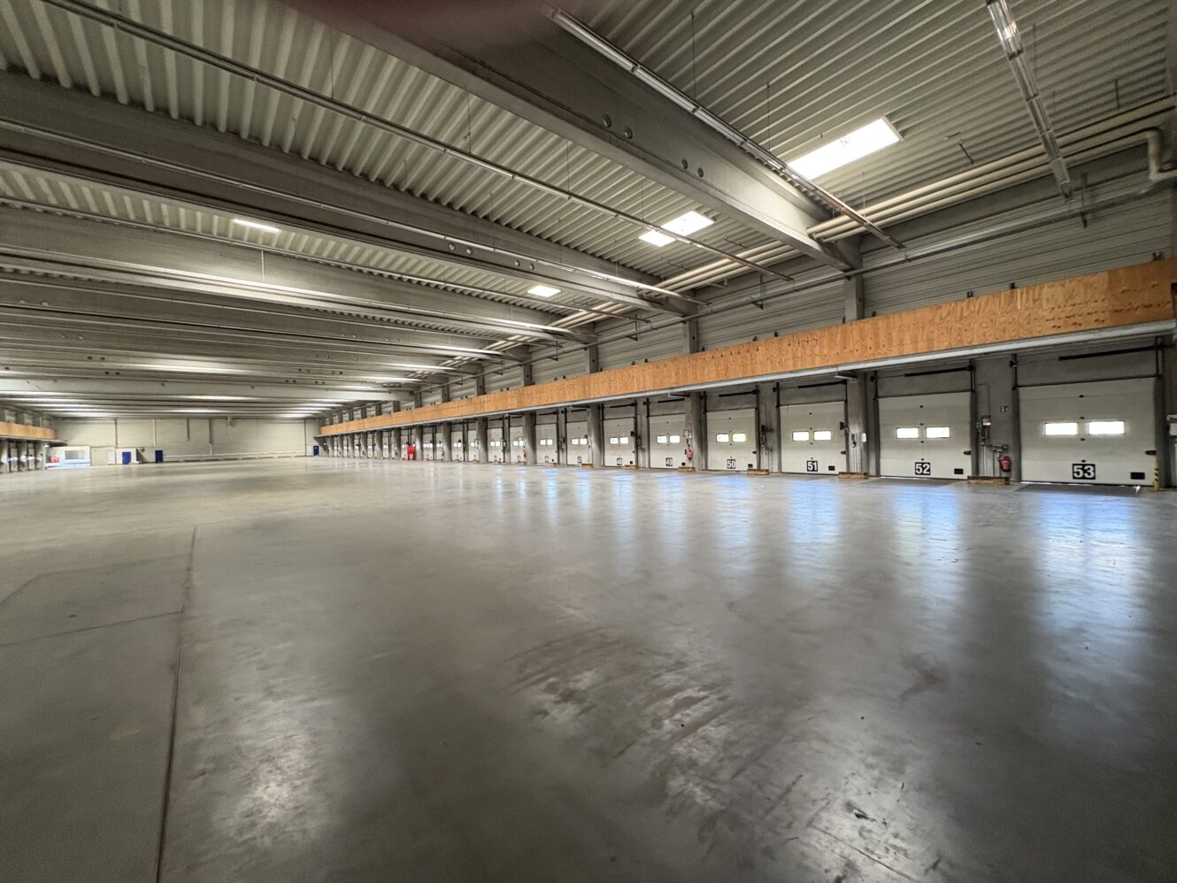 Weitwinkelaufnahme des Innenraums eines leeren Lagerhauses mit mehreren Laderampentüren und Deckenleuchten, die den Betonboden beleuchten.