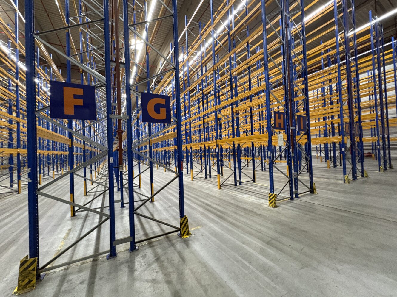 Leere Industriehalle mit blauen und gelben Metallregalen, Betonboden und Schildern mit den Buchstaben F, G und H.