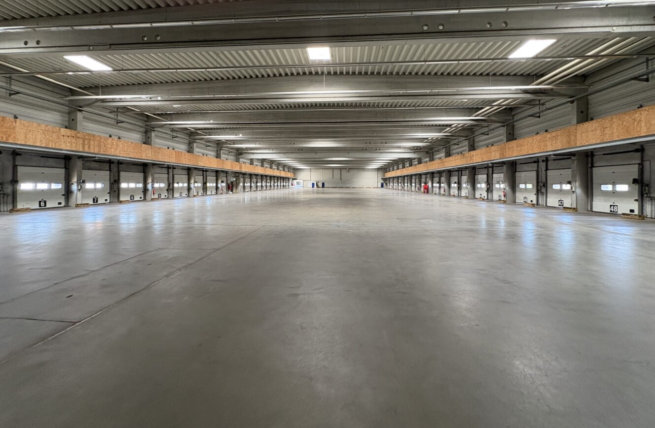 Innenansicht einer leeren Industriehalle mit glattem Betonboden, hoher Decke und mehreren Ladetoren entlang der Wände, aufgenommen aus der Mitte des Raums.