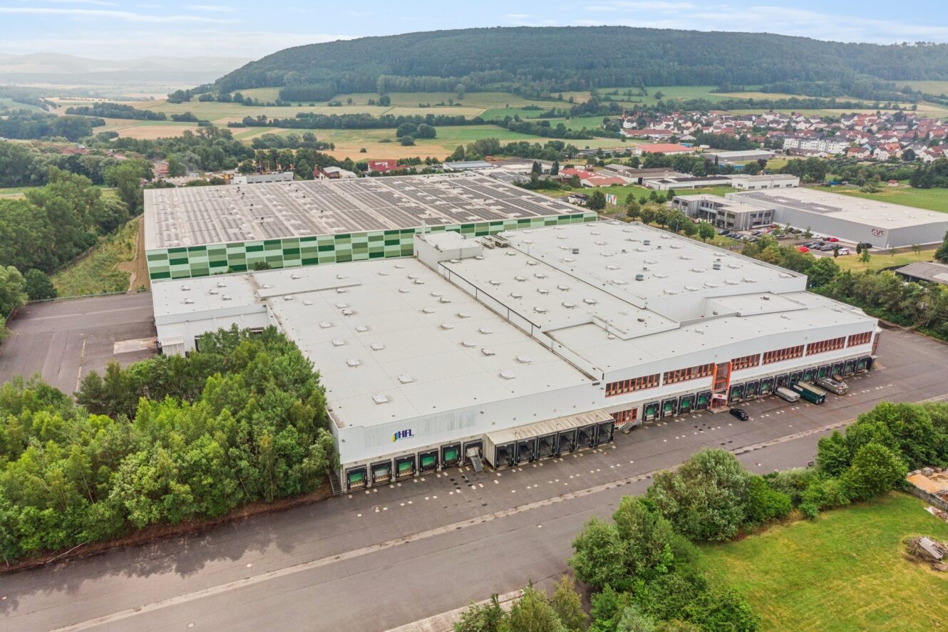 Großes Industrie-Lagerhaus mit mehreren Laderampen, umgeben von Bäumen und offenem Gelände, im Hintergrund eine kleine Stadt und Hügel.