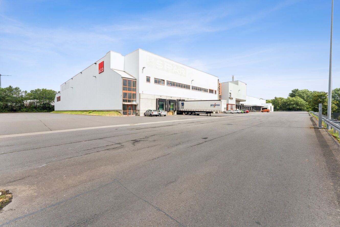 Großes weißes Lagerhaus mit Laderampen, ein geparkter Sattelzug, ein weißer Transporter in der Nähe, ein leerer gepflasterter Platz und umgebendes Grün unter blauem Himmel.