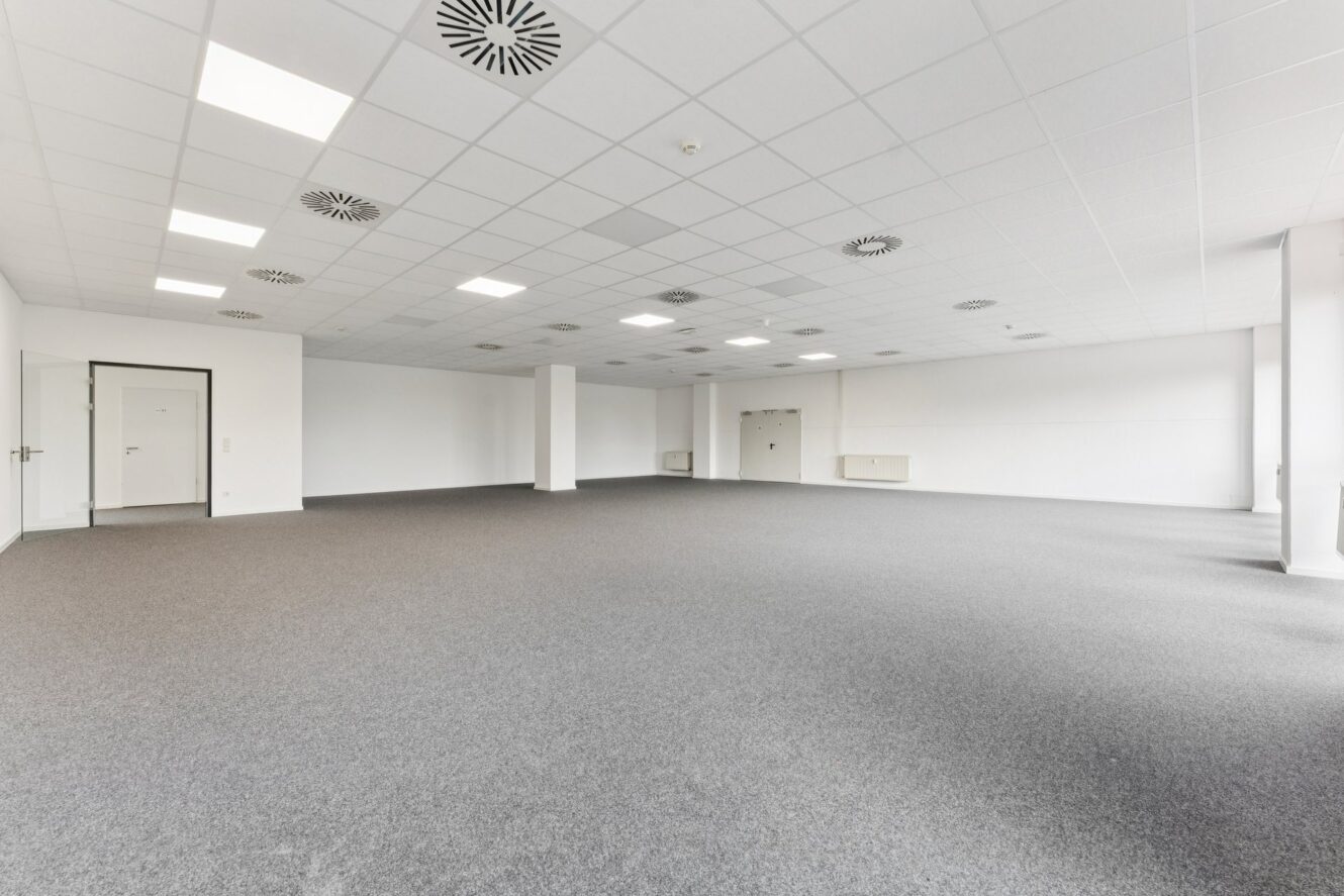 Geräumiger, leerer Büroraum mit weißen Wänden, grauem Teppichboden, abgehängter Decke mit Leuchten und Lüftungsgittern.