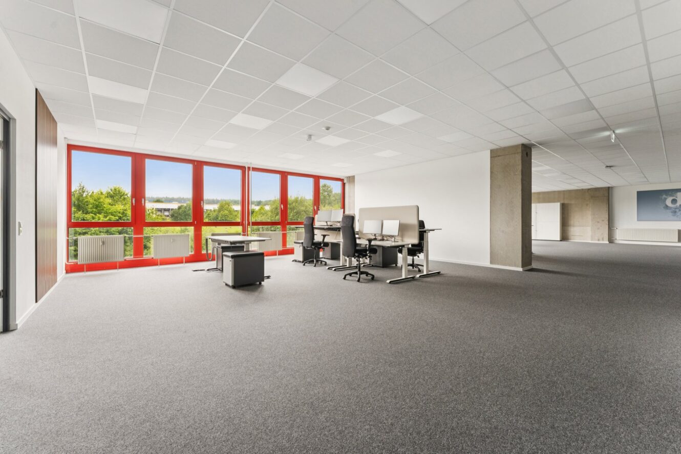 Ein modernes, geräumiges Büro mit grauem Teppichboden, mehreren Schreibtischen und Stühlen, großen Fenstern mit roten Rahmen und Blick auf die Bäume draußen. Der Raum ist größtenteils leer und wird von Tageslicht gut beleuchtet.